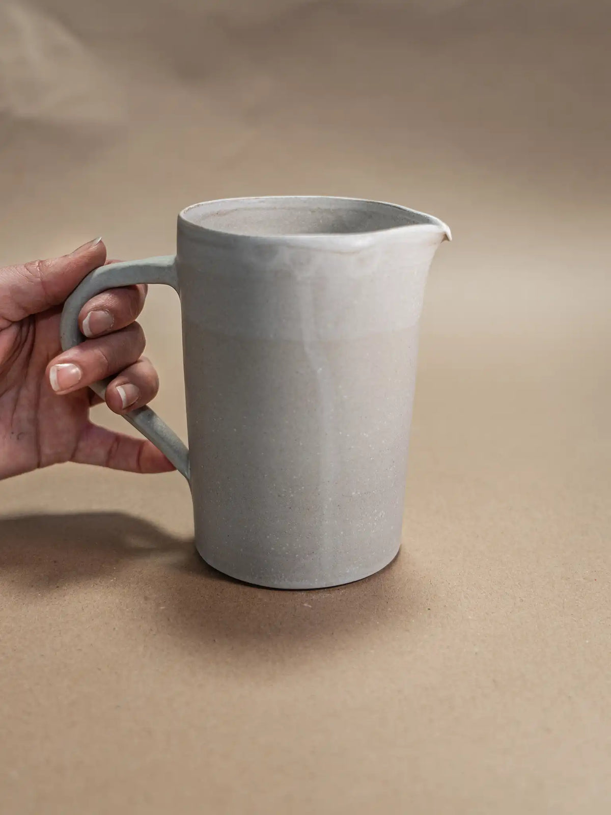 Een minimalistische handgemaakte schenkkan in de kleur taupe van Iris Floor Ceramics keramiek. De kan heeft een strak handvat en een subtiel glazuurverloop aan de bovenzijde.