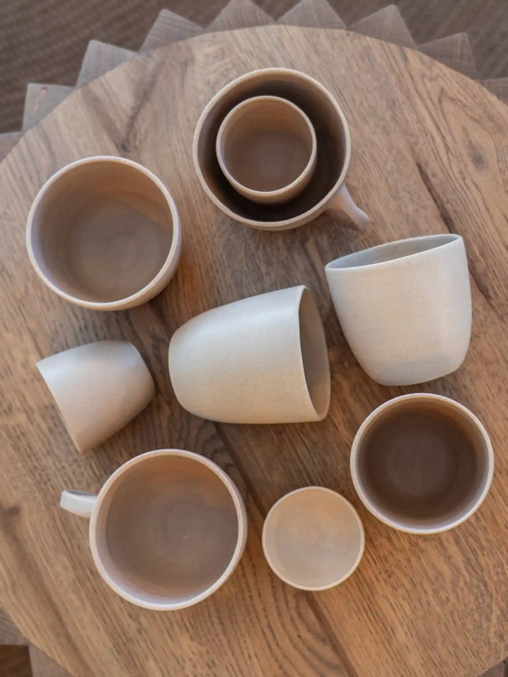Handgemaakte keramieken mokken en bekers in verschillende maten en aardtinten, bovenaanzicht op ronde houten tafel — Iris Floor Ceramics