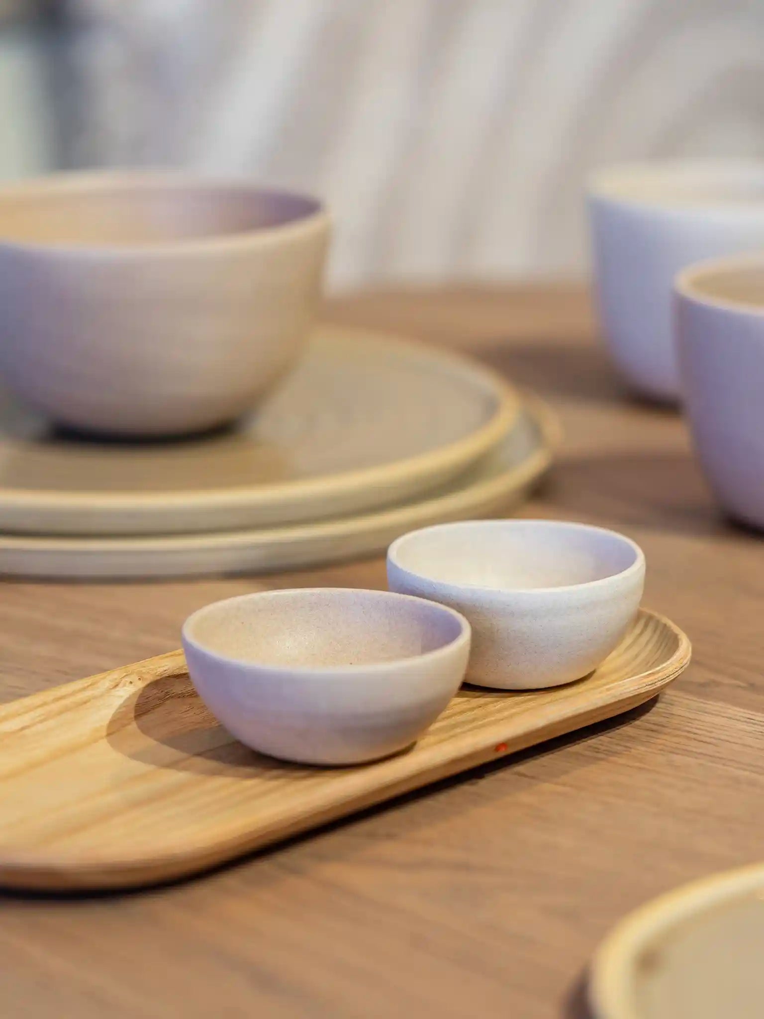 Twee kleine handgemaakte keramieken schaaltjes in lila-wit glazuur op een houten dienblad, met borden en bekers op de achtergrond — Iris Floor Ceramics