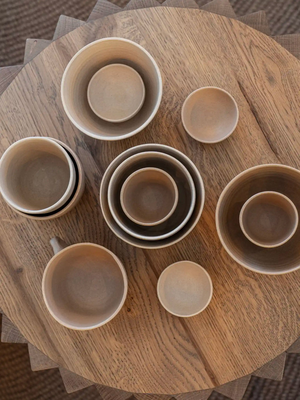 Handgemaakte keramieken serviesset in aardtinten — kommen, mokken en schaaltjes in verschillende maten op een ronde houten tafel, door Iris Floor Ceramics