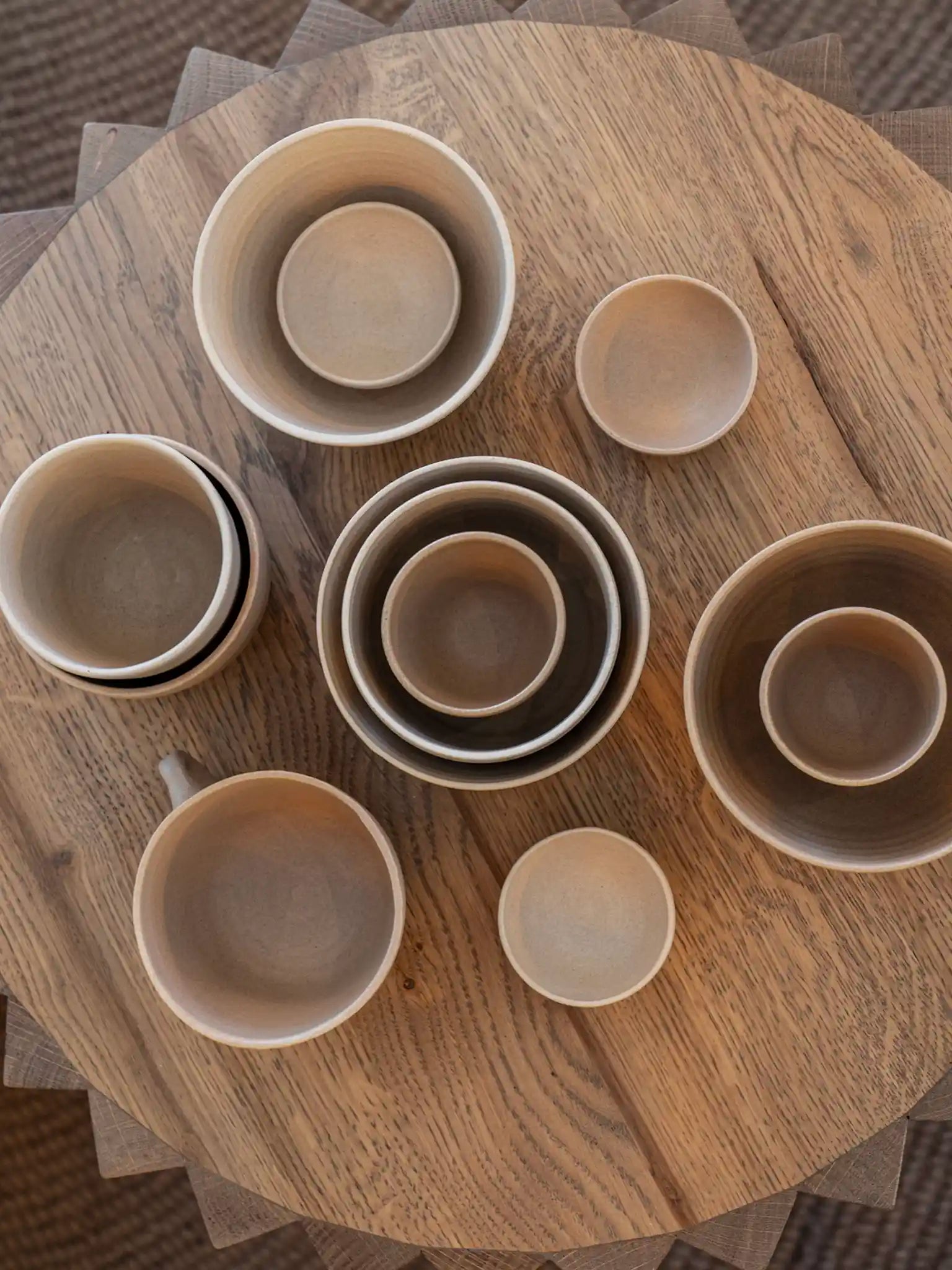 Handgemaakte keramieken serviesset in aardtinten — kommen, mokken en schaaltjes in verschillende maten op een ronde houten tafel, door Iris Floor Ceramics